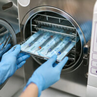 Technician in blue latex gloves is inserting sealed dental probes inside an autoclave sterilizer. Closeup horizontal photo.